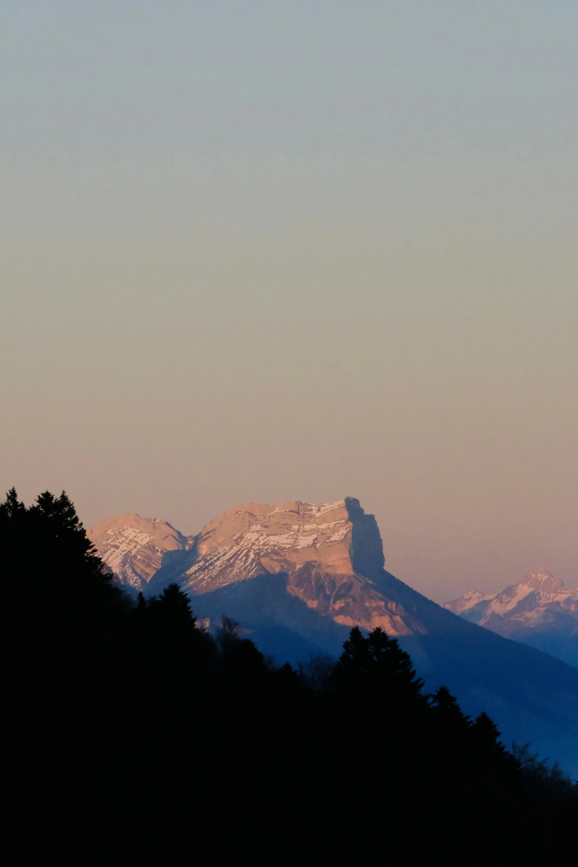 A sunlit rocky mountain ridge behind dark silhouetted pine hills under a soft pink-and-blue dusk haze.