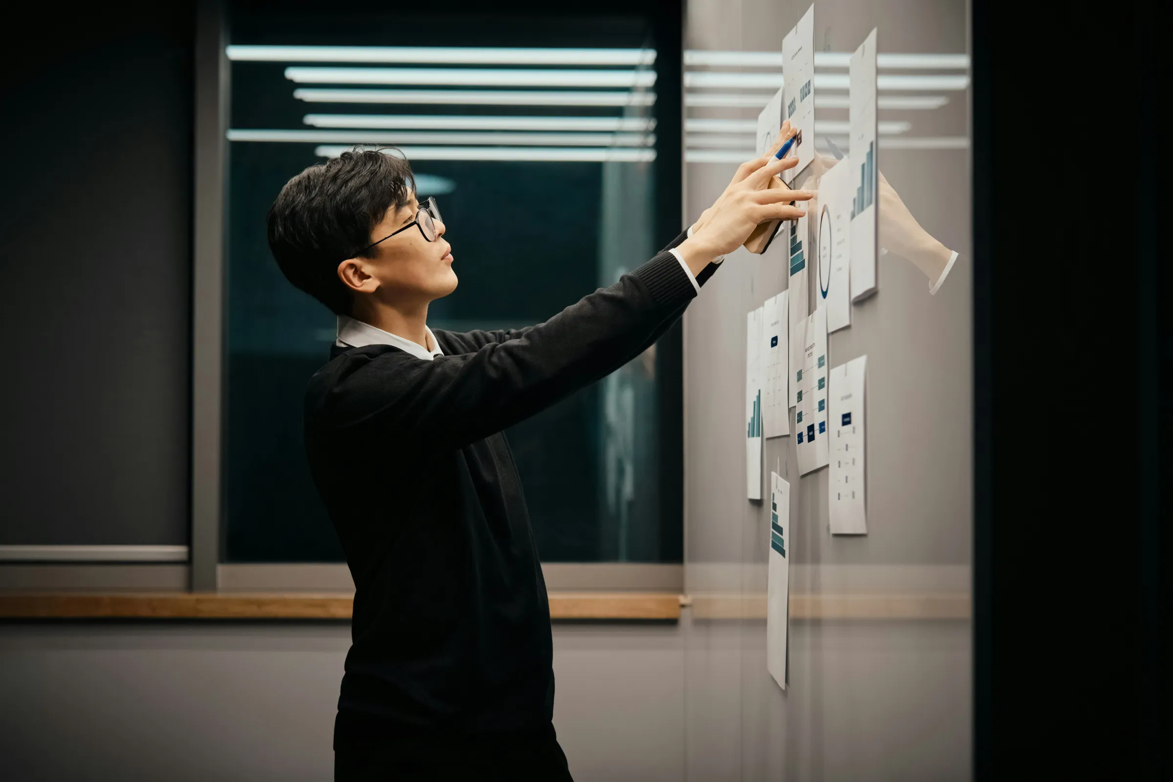 A man in a dark sweater and glasses reaches up to pin printed charts and dashboards onto a glass wall in a dim room.