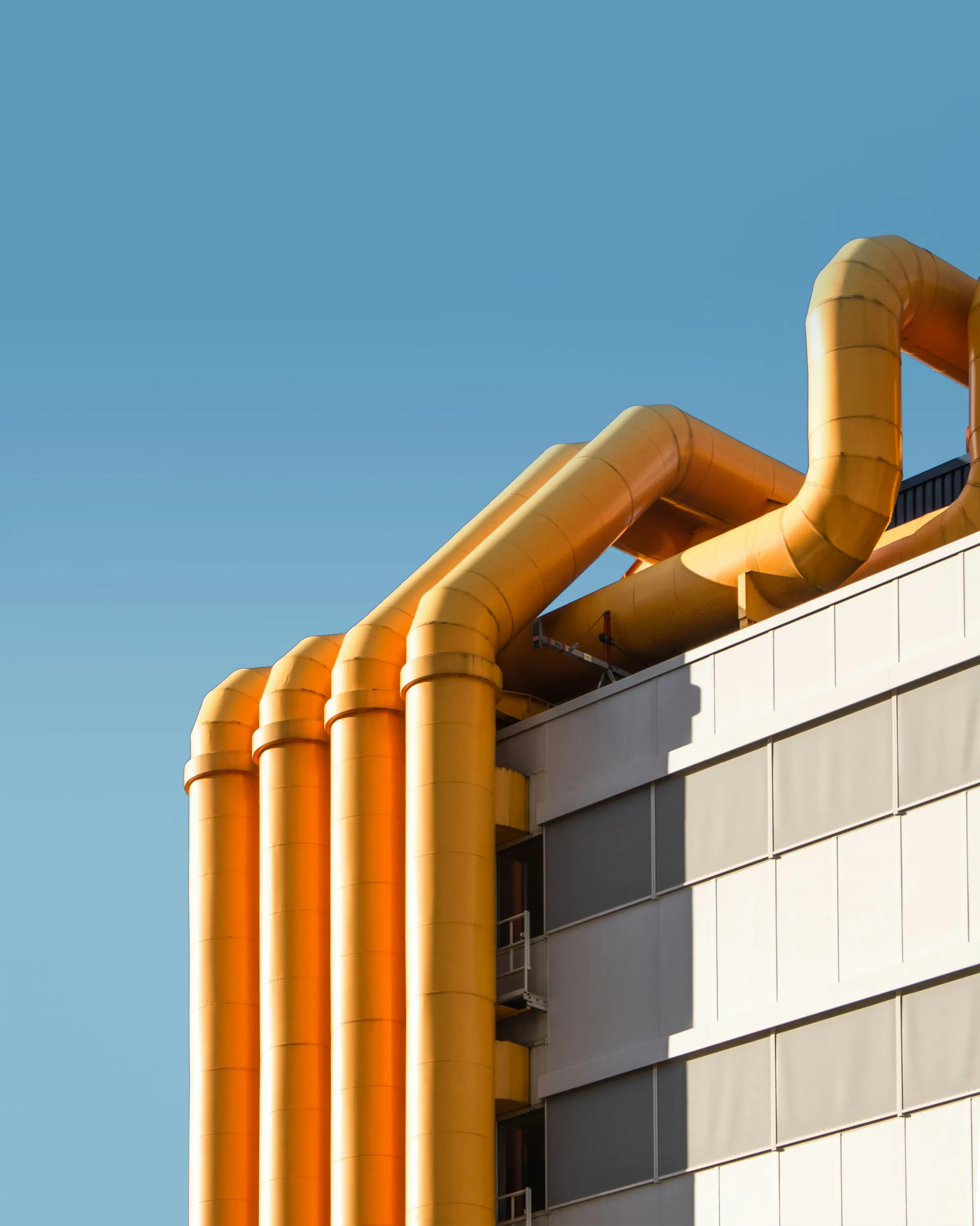 Gold industrial ducting pipes curving across the top corner of a grey-panelled building against a clear blue sky.