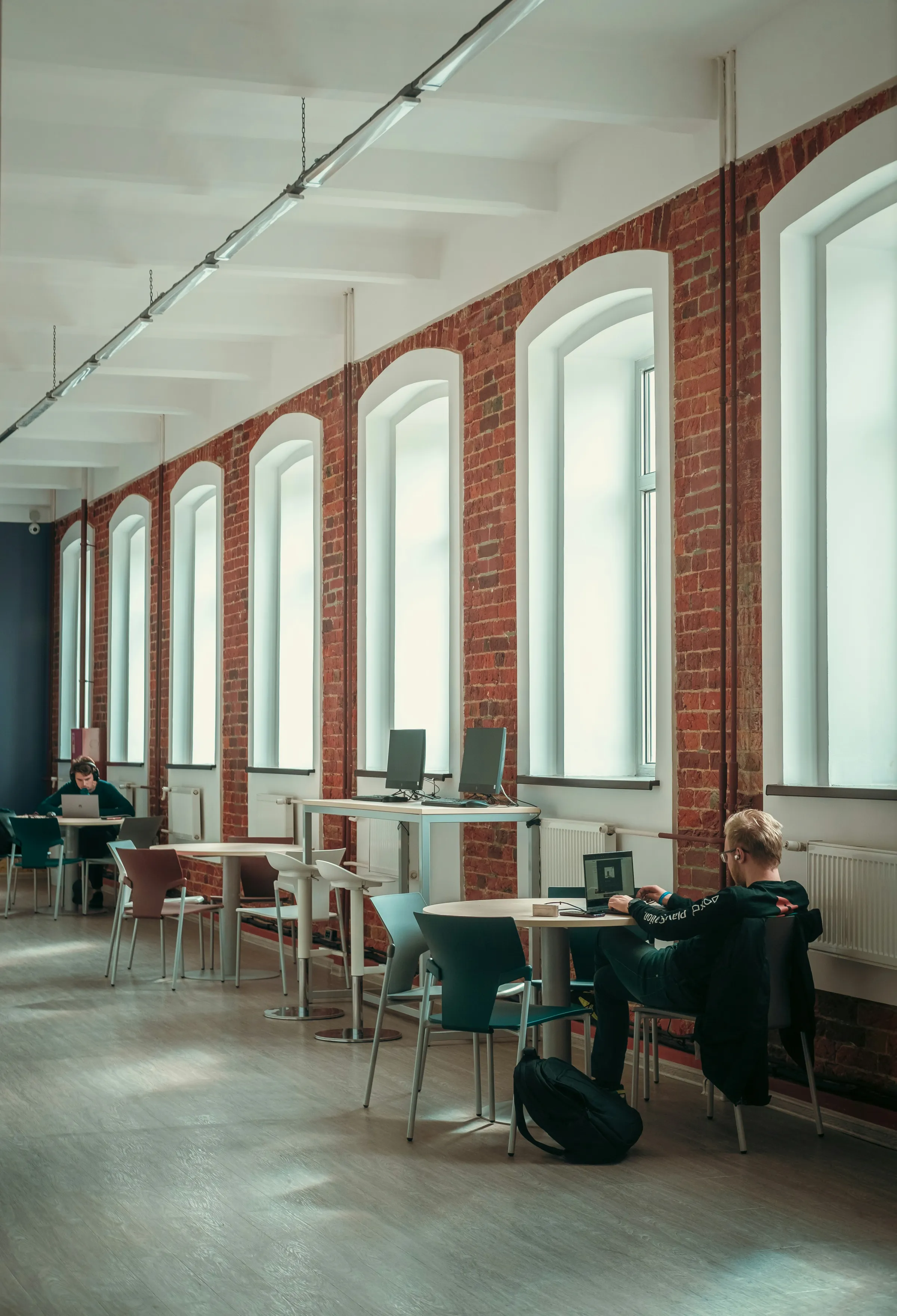 A long converted-warehouse study space with tall arched brick windows, round tables, and people working on laptops.
