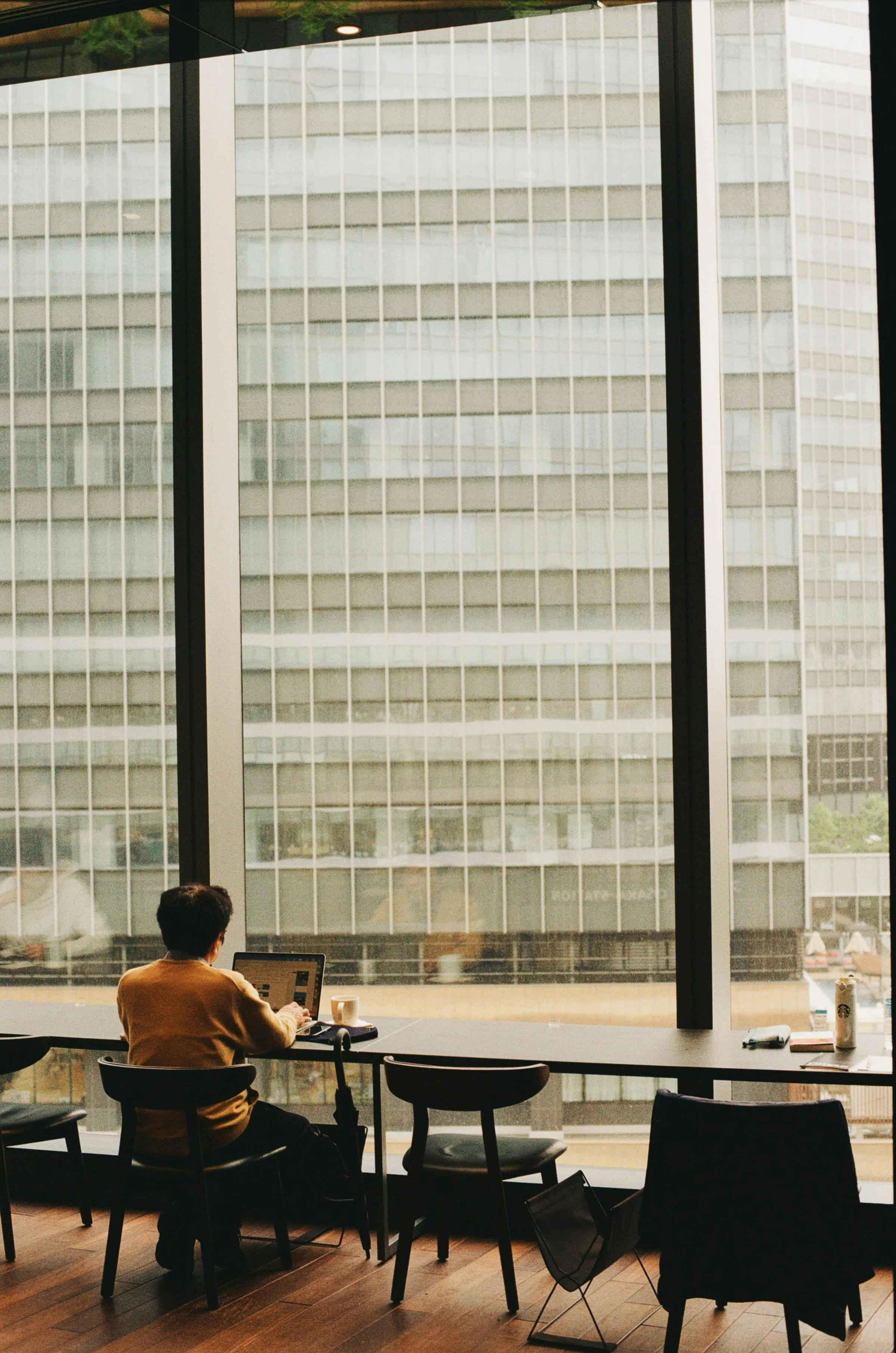 A person in a mustard cardigan works on a laptop at a long counter facing a large window that overlooks a grey office tower.