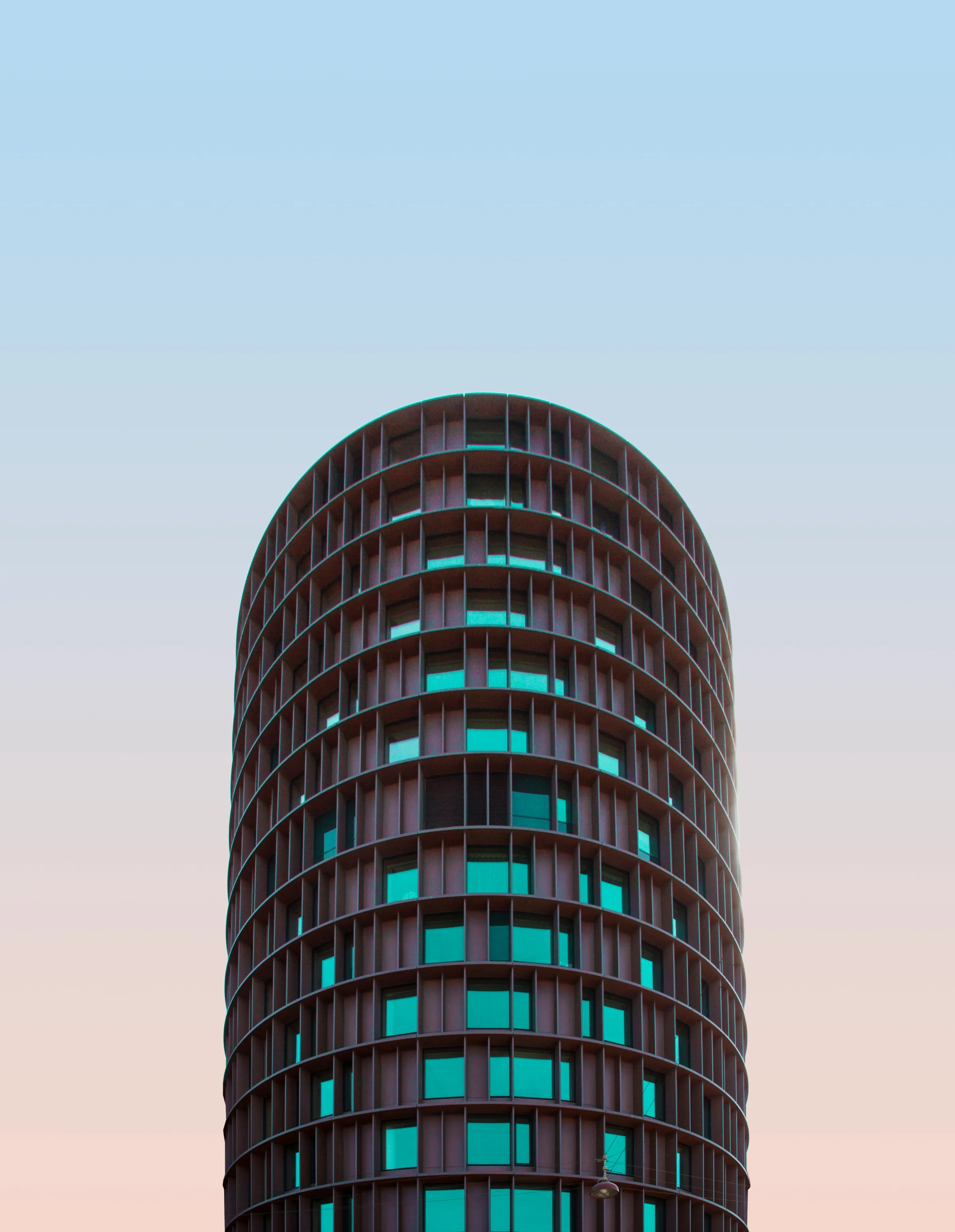 A rounded-top cylindrical tower with a dark red facade and glowing teal windows against a pastel sky.