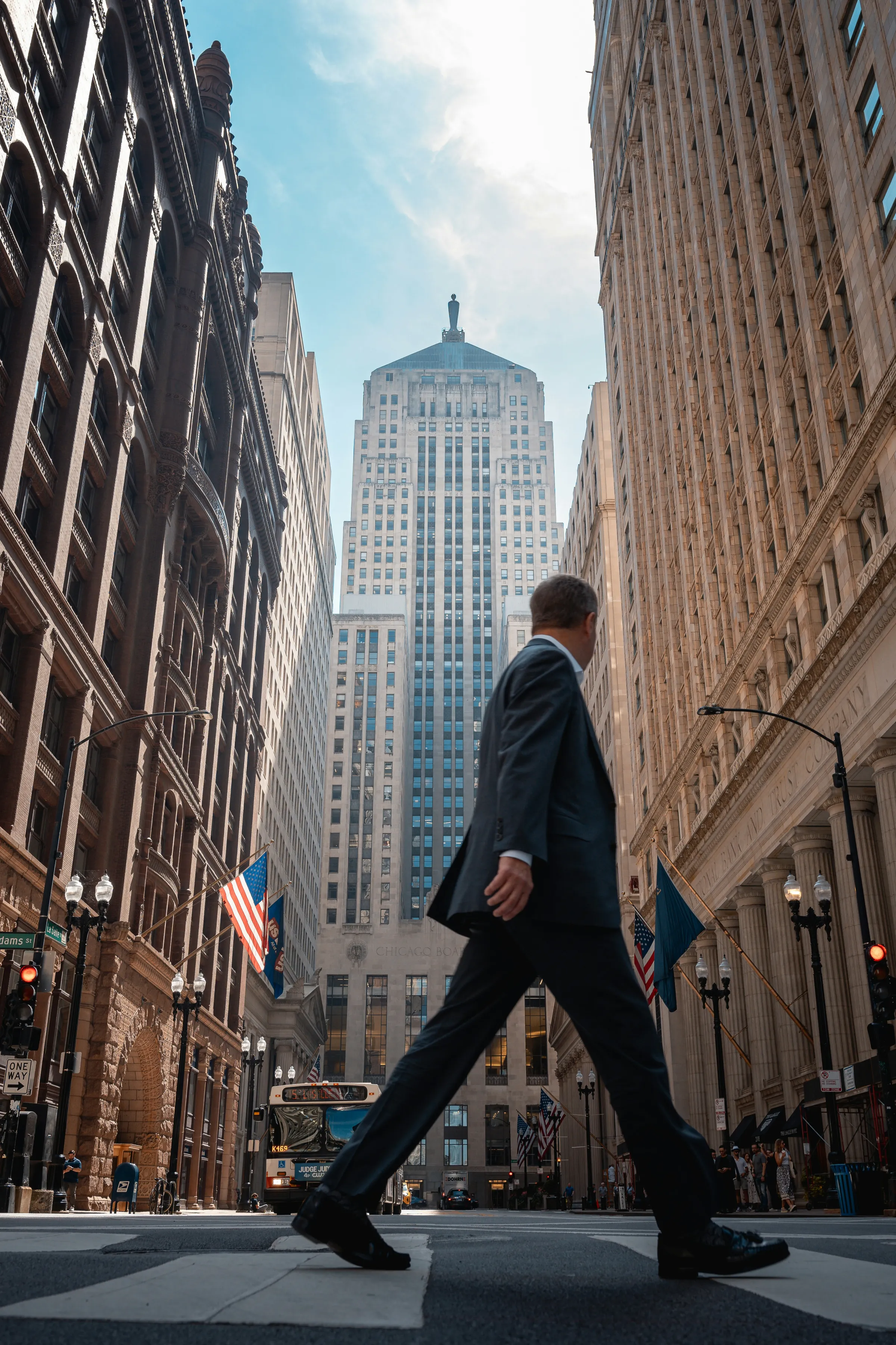 A low-angle view of a suited man striding across a crosswalk with the Chicago Board of Trade tower rising between office buildings.