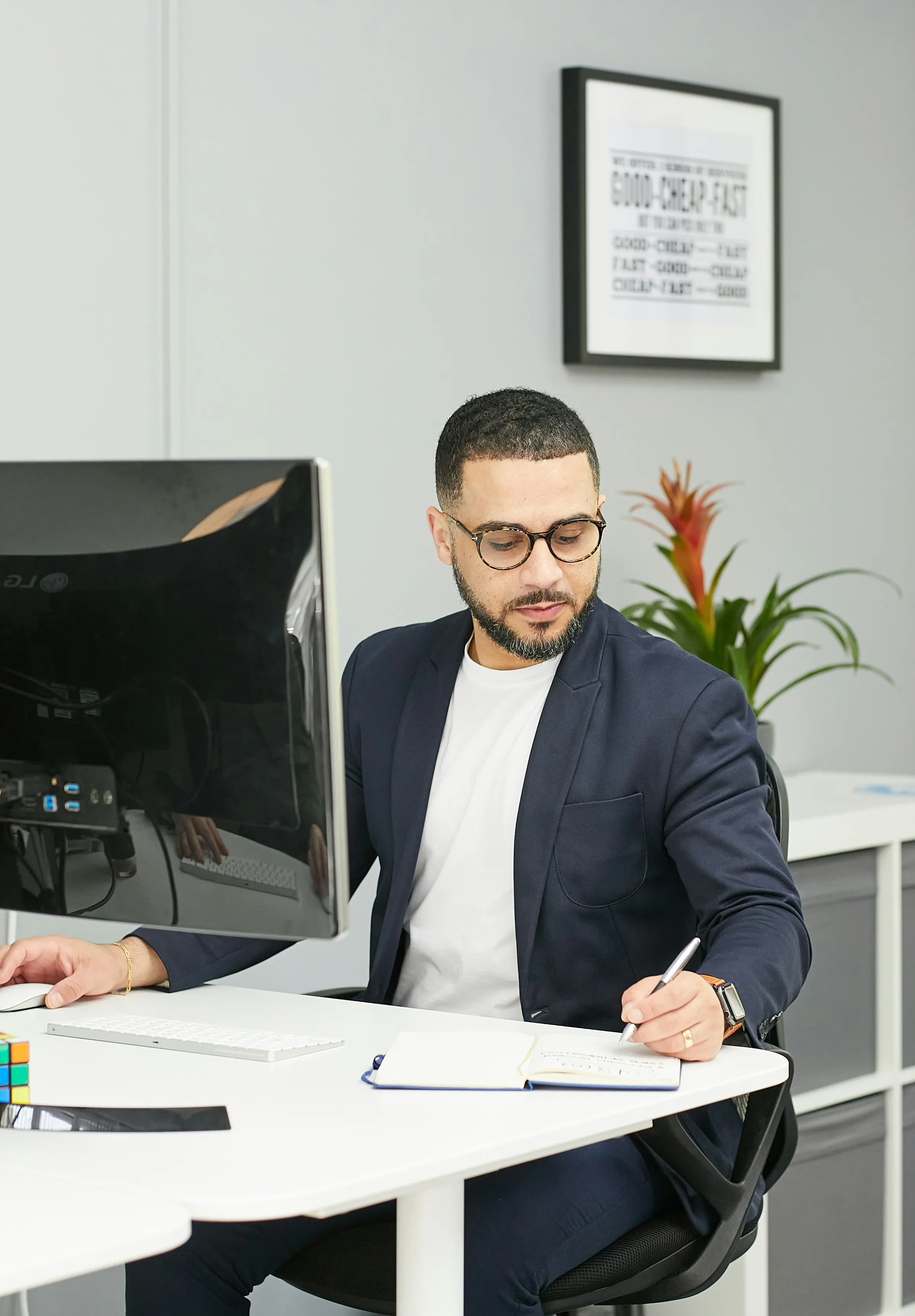 A man in a navy blazer and glasses writes in a notebook at a white desk with a monitor and a framed 'good-cheap-fast' print behind.