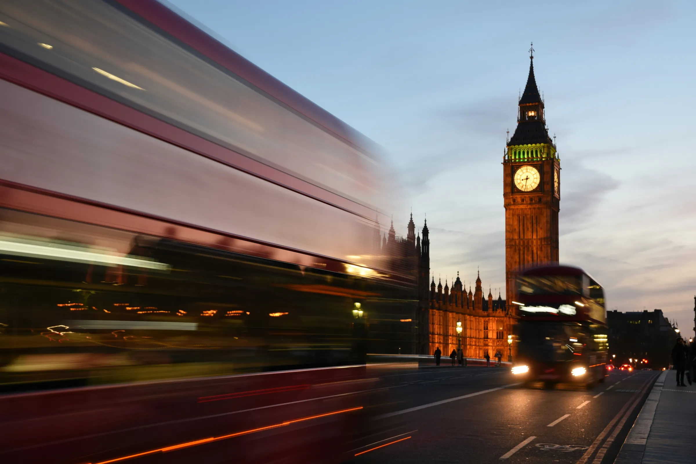 Big Ben lit at dusk with a motion-blurred red double-decker bus crossing Westminster Bridge.