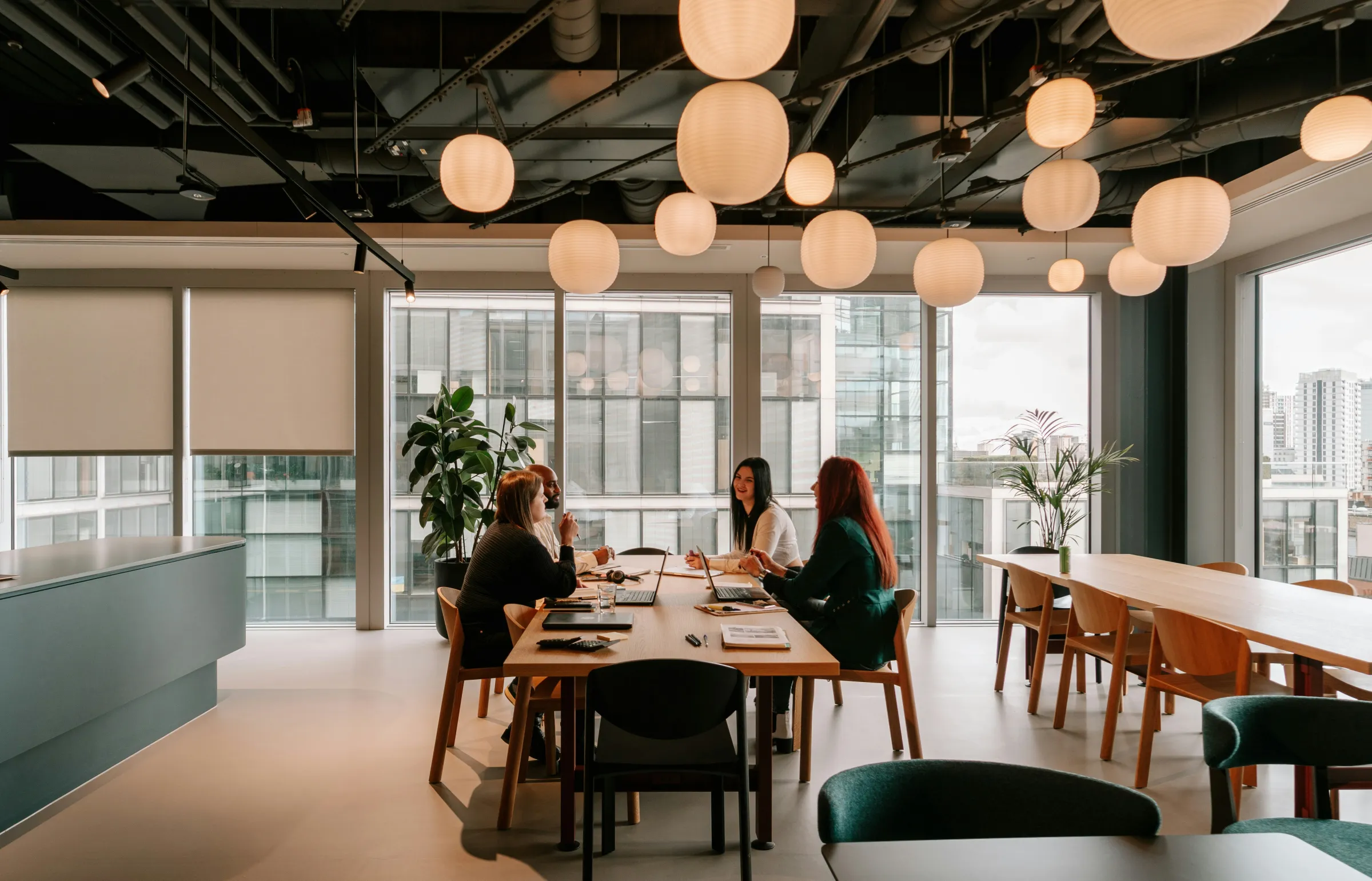 Four colleagues meeting at a wooden table in a modern coworking space lit by clustered paper-lantern pendants.