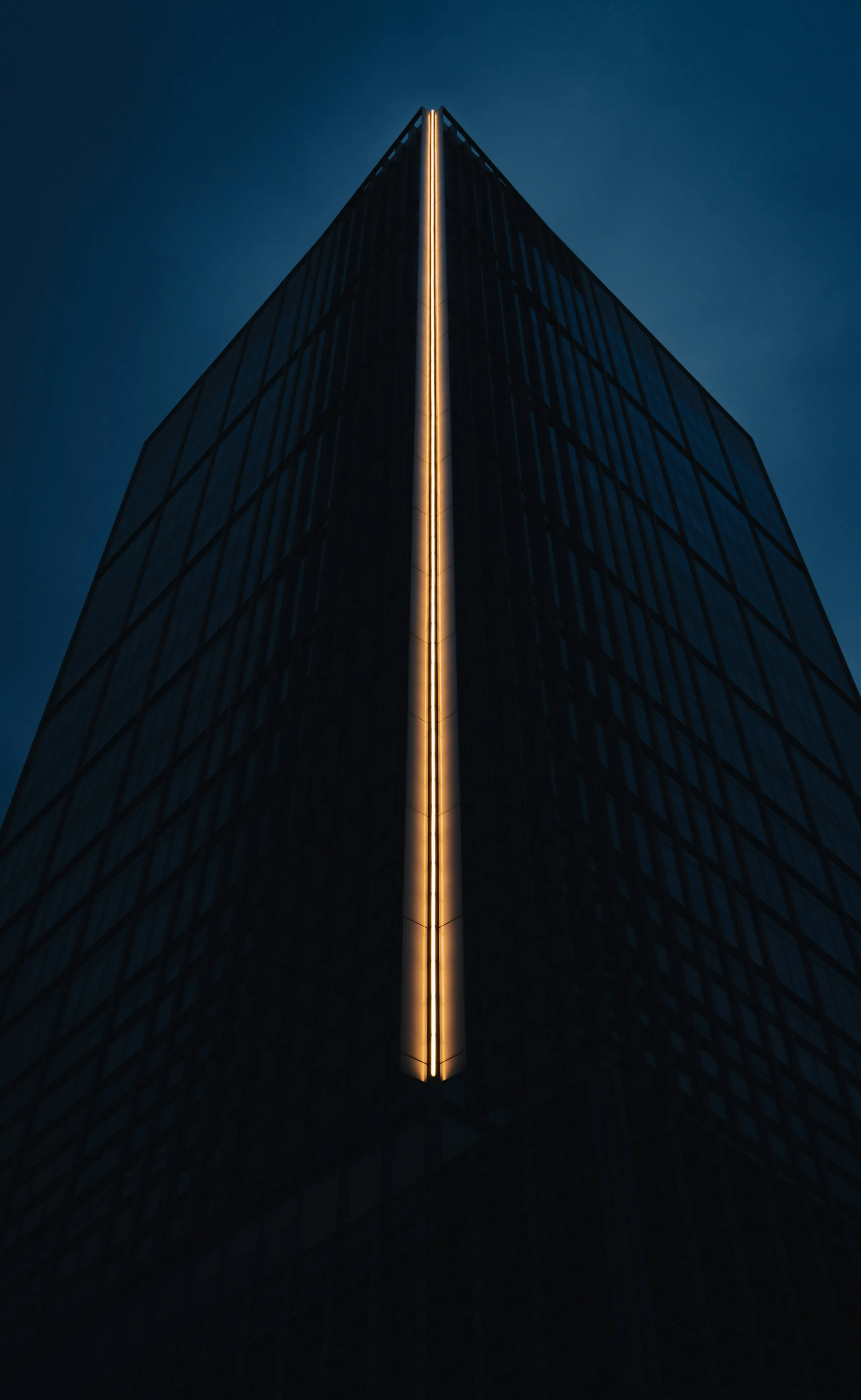 A near-black glass skyscraper viewed from below at dusk, a glowing gold vertical light seam running up its corner edge against a deep blue sky.