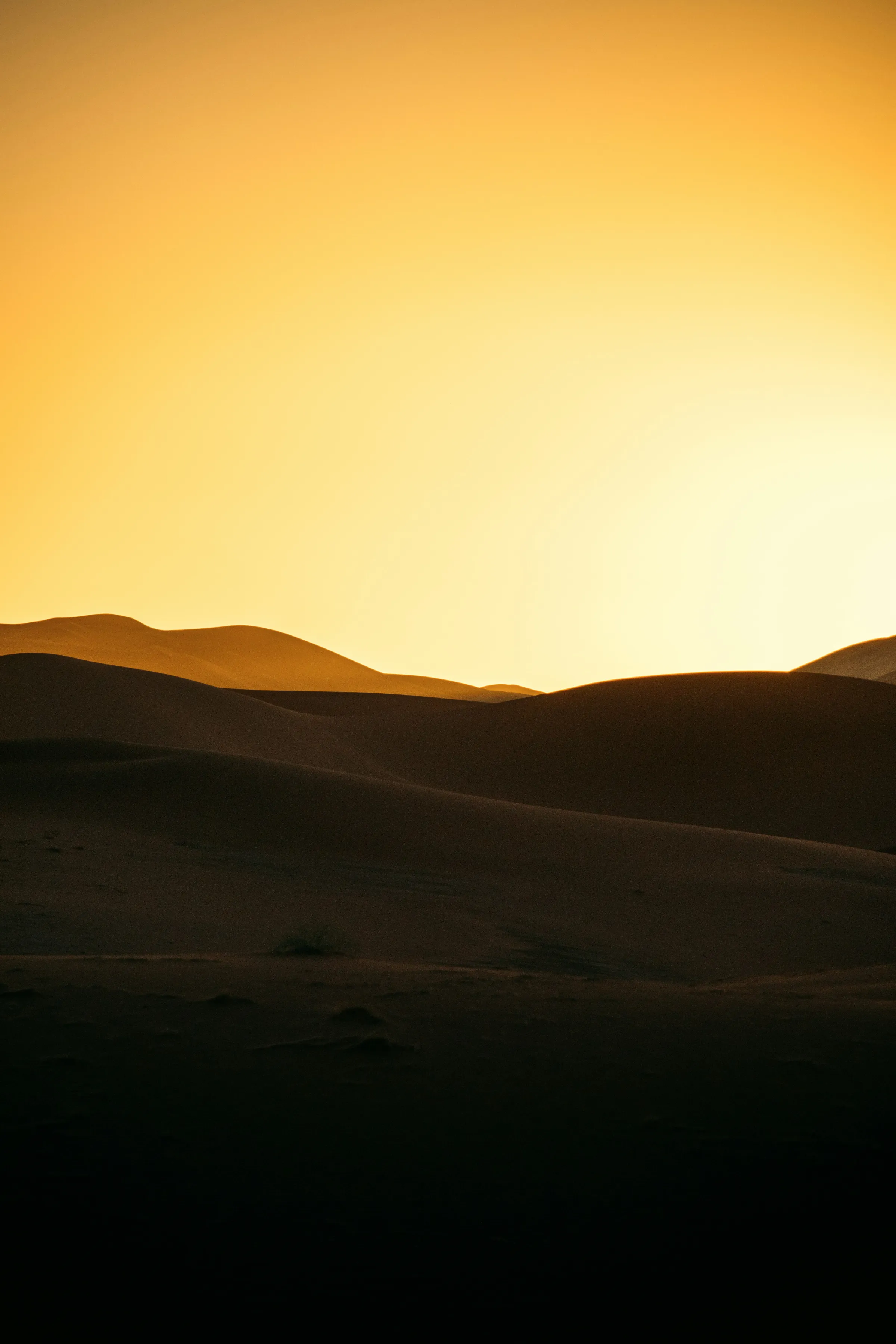 Rolling sand dunes silhouetted against a glowing golden-yellow desert sky at sunset.