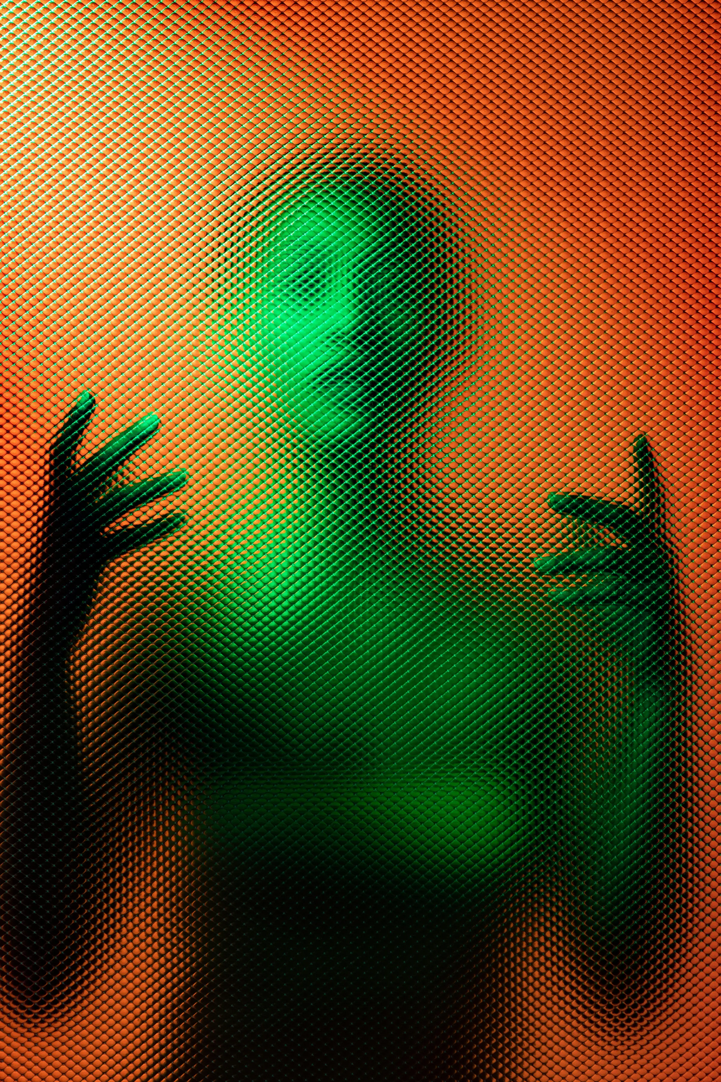A face and hands press against taut mesh fabric, lit green at centre against a surrounding orange glow.