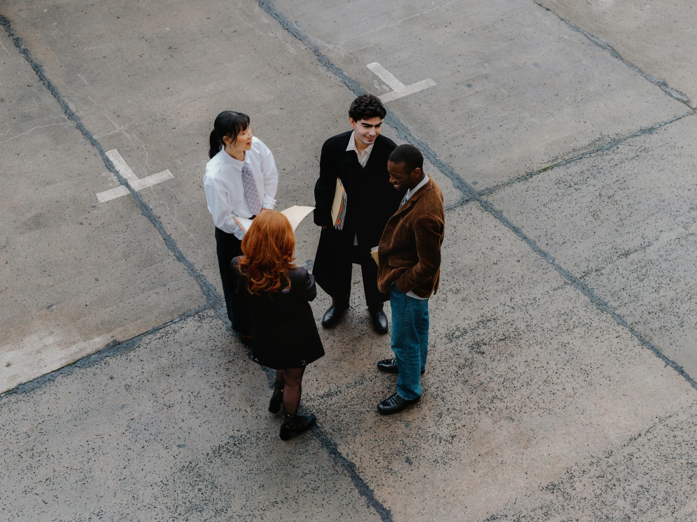 Overhead view of four business people in smart-casual dress standing talking on a cracked concrete lot marked with white cross symbols.