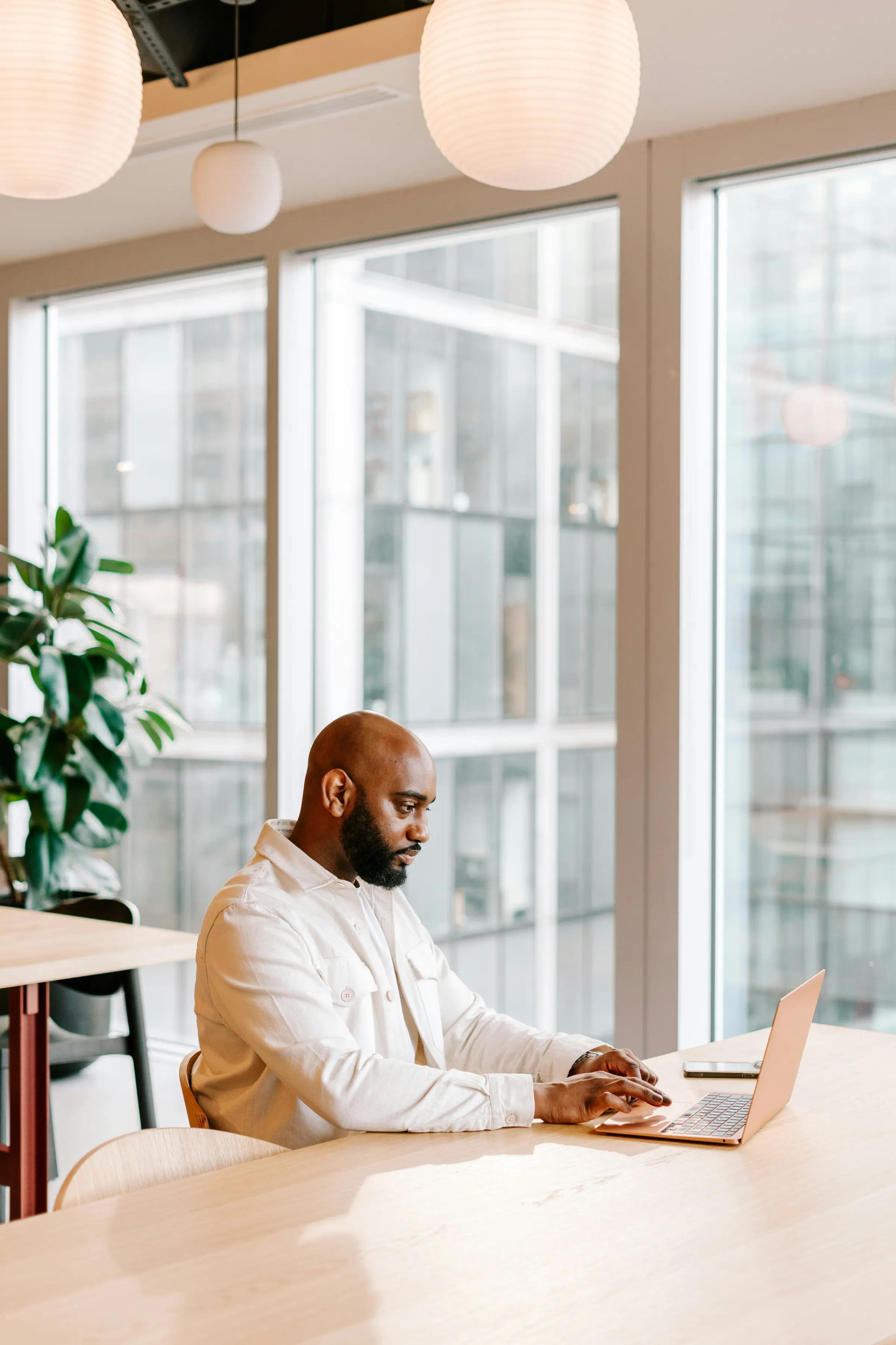 A bearded man in a cream jacket works on a laptop at a wooden table beside a bright floor-to-ceiling window.