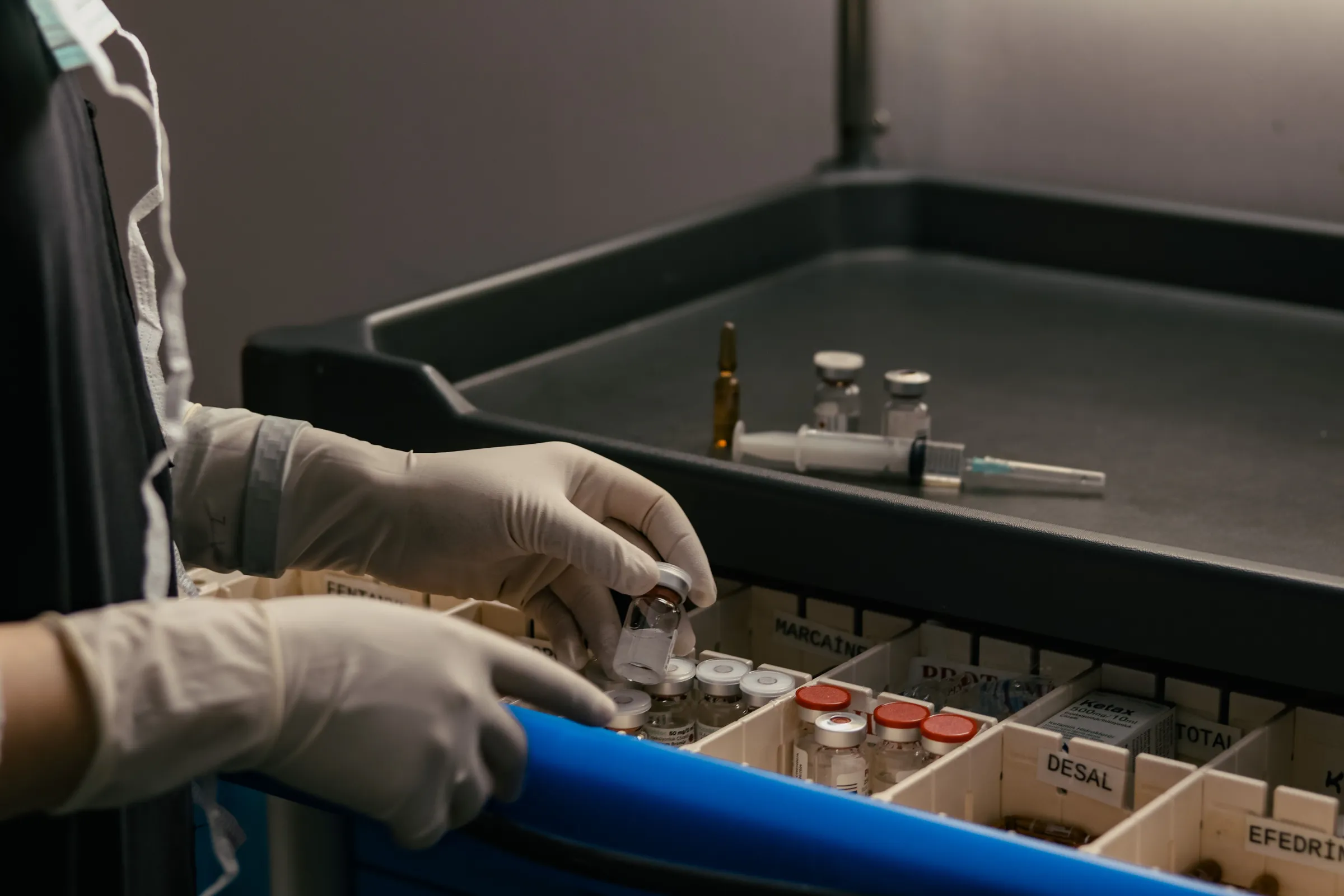 Gloved hands selecting a labelled medication vial from a compartmentalised anaesthetic drug cart, with a syringe and vials on a tray behind.