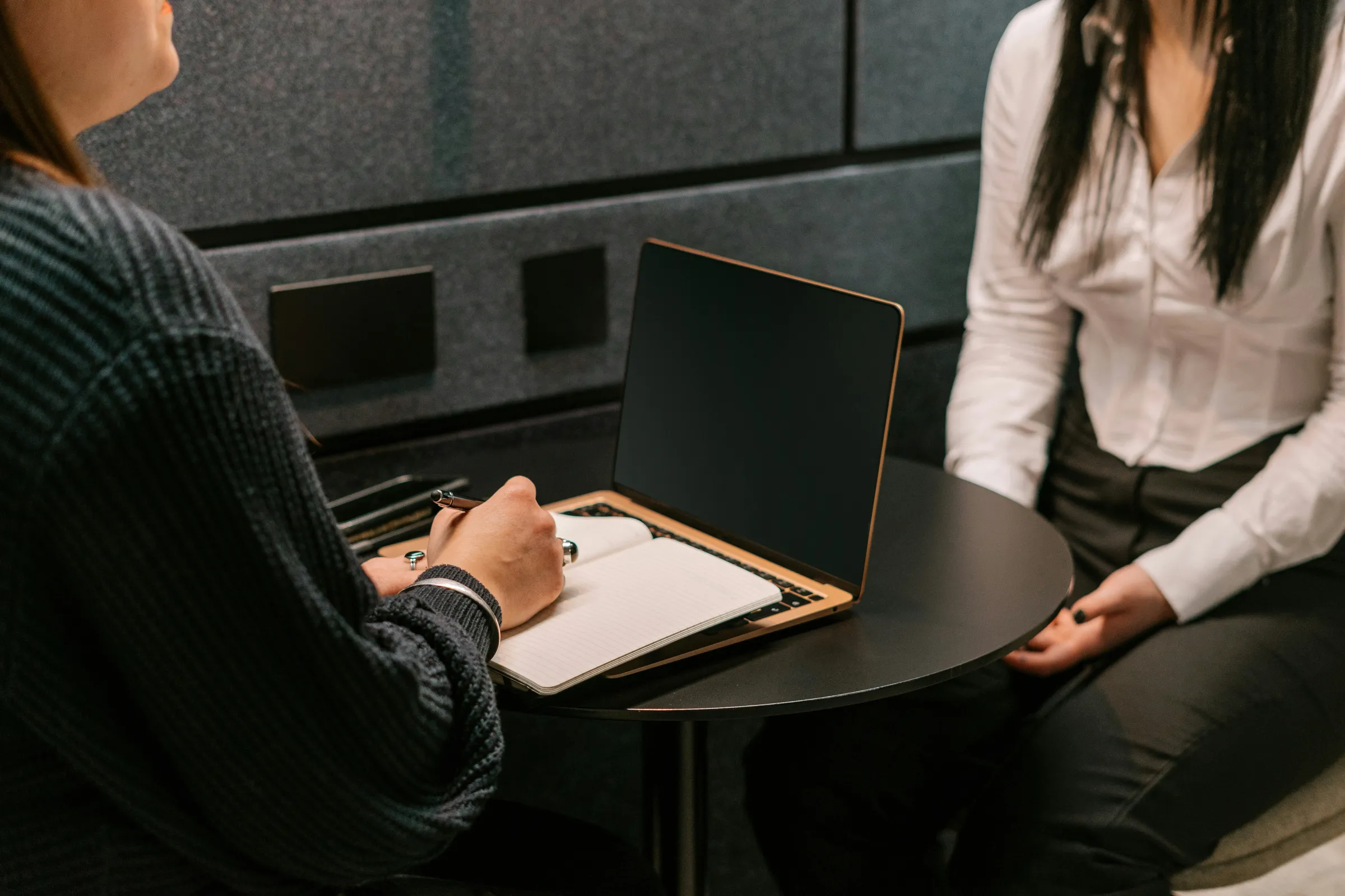 Two women in business dress meet across a small round table with a laptop in a dark acoustic booth.