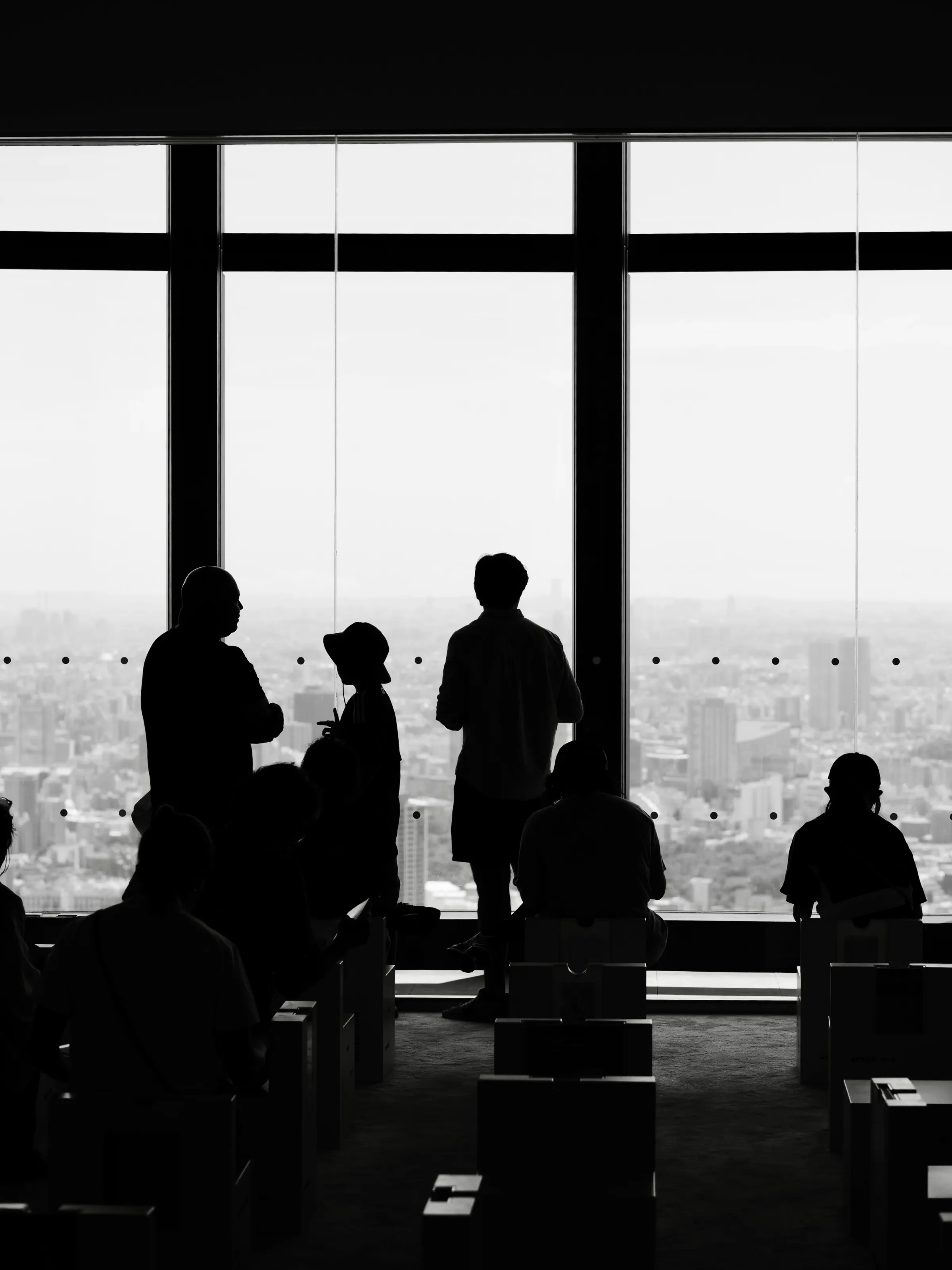 Black-and-white silhouettes of people standing and sitting at a floor-to-ceiling window overlooking a hazy city skyline.