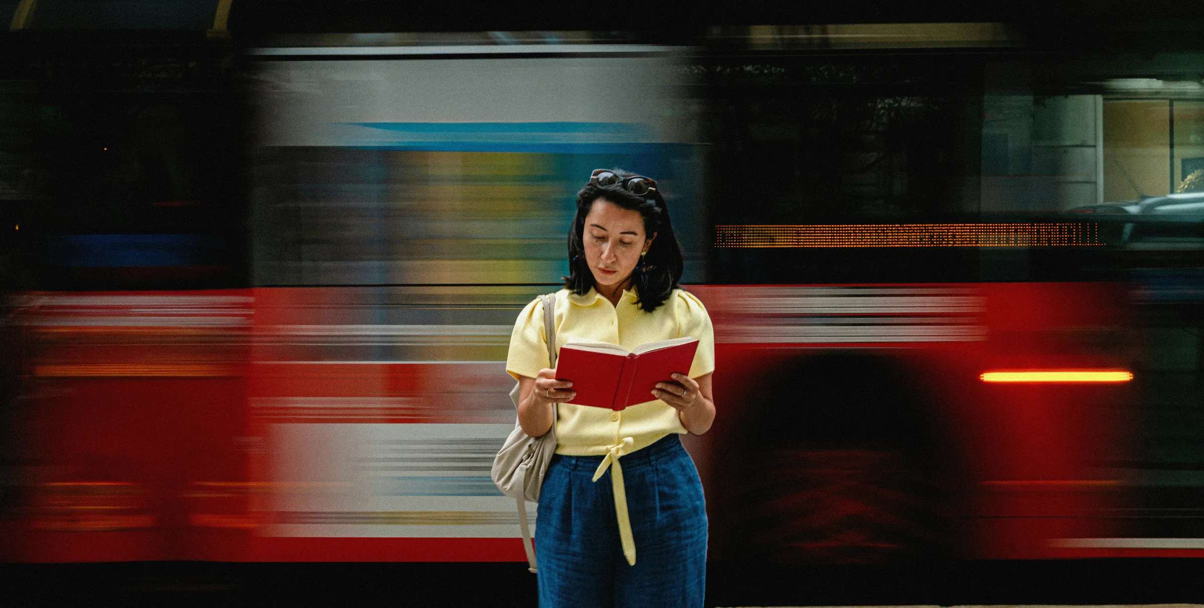 A woman in a yellow blouse reads a red book while a red bus streaks past behind her in motion blur.