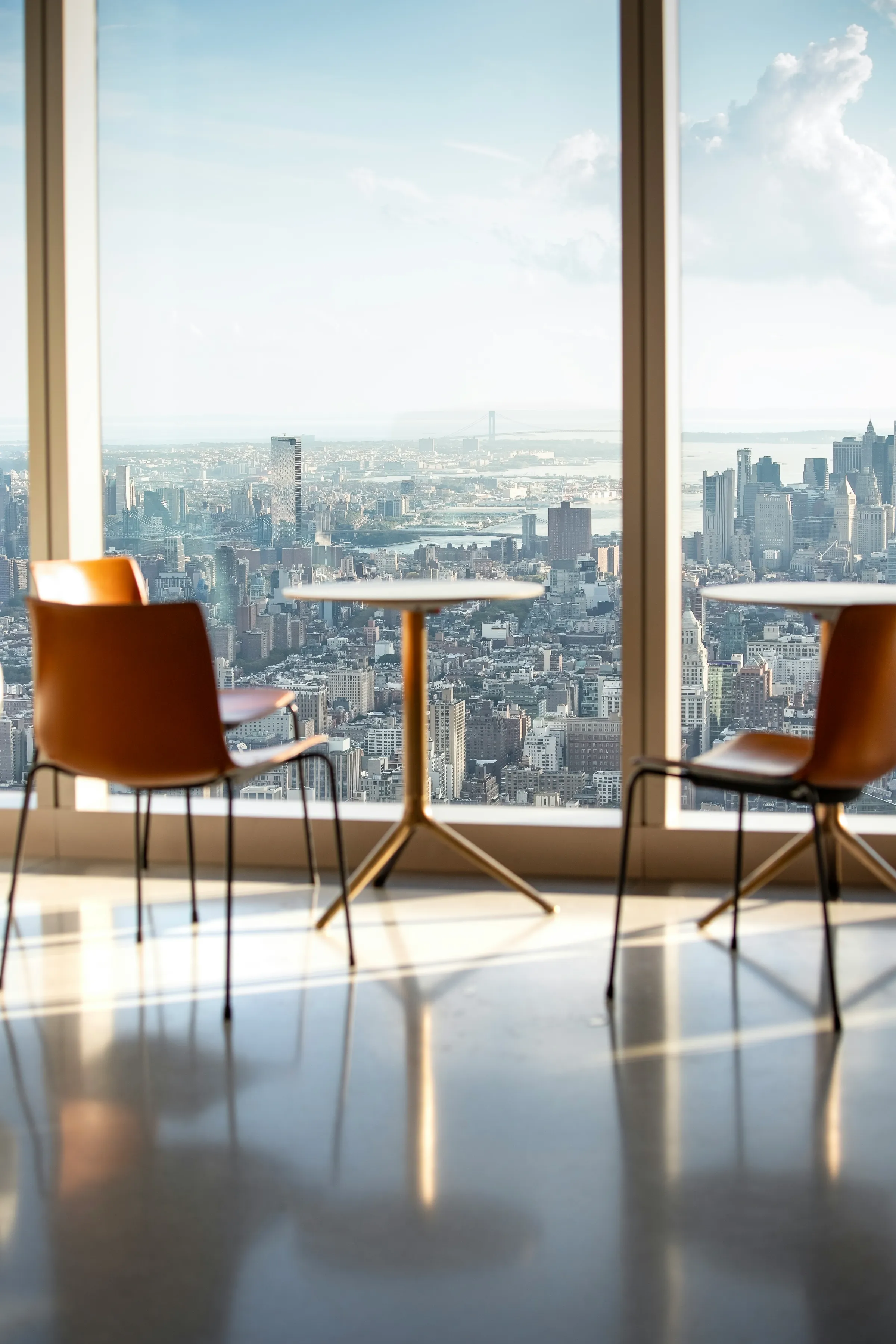 Two tan leather chairs and small round tables by a floor-to-ceiling window looking out over a hazy Manhattan skyline.