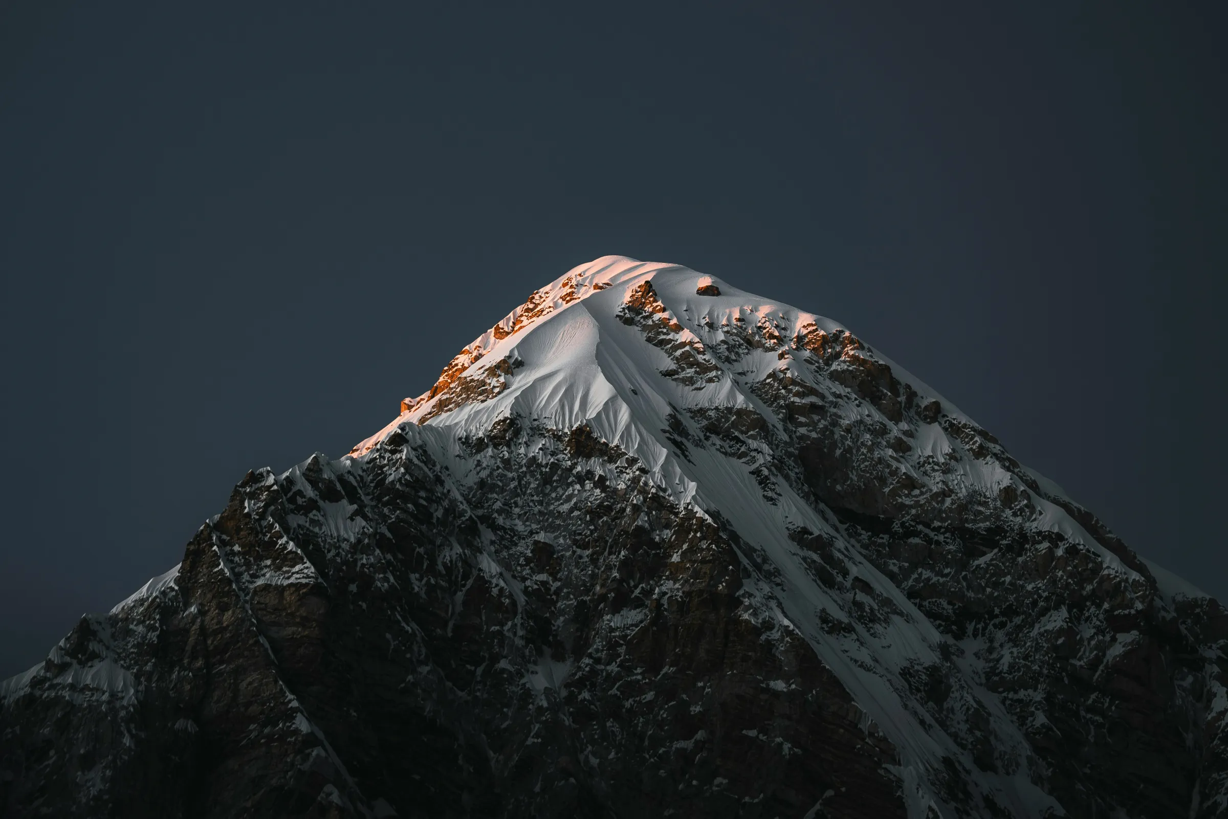 A snow-covered rocky mountain summit catching faint warm light against a near-black night sky.