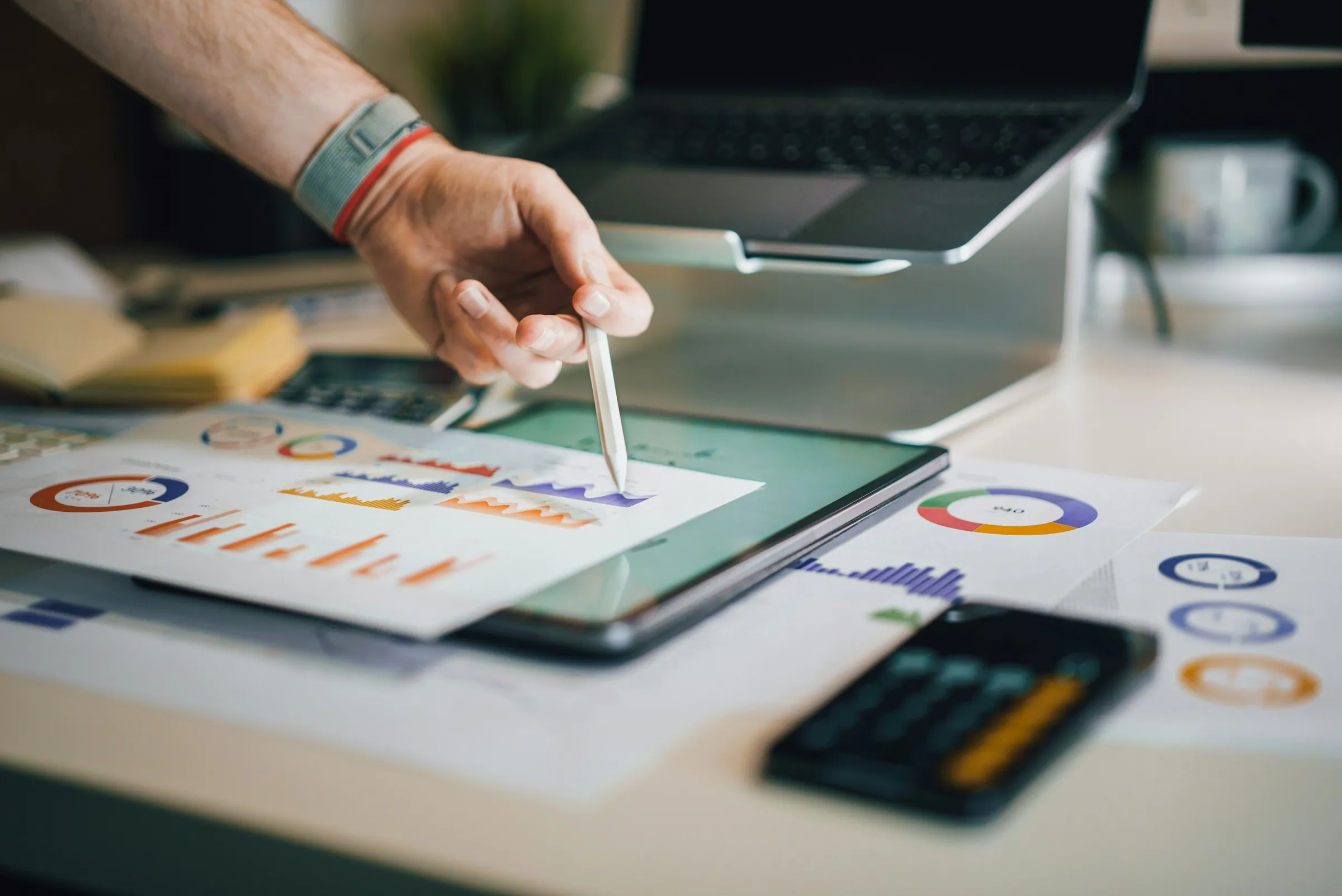 A hand with a white stylus points at a tablet on a desk surrounded by printed charts, pie graphs, a calculator, and a laptop.