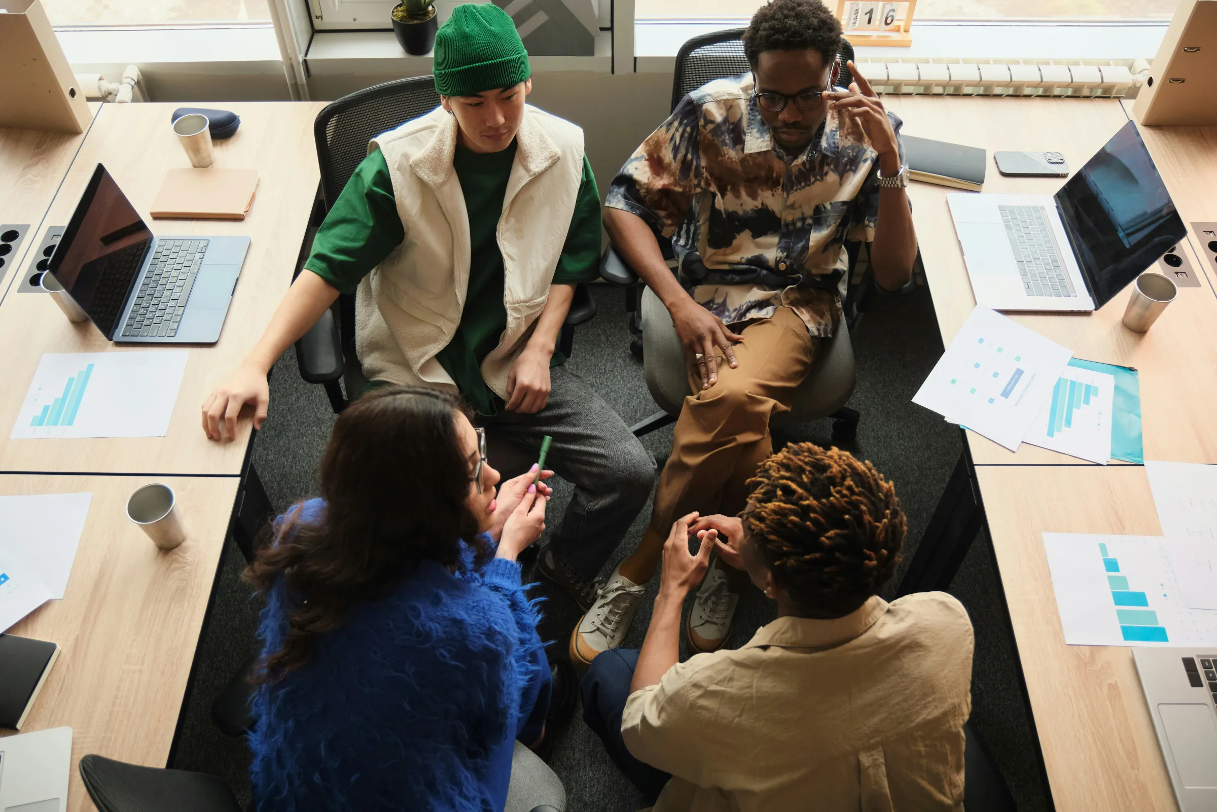Overhead view of four colleagues gathered around desks scattered with laptops and printed bar-chart reports.