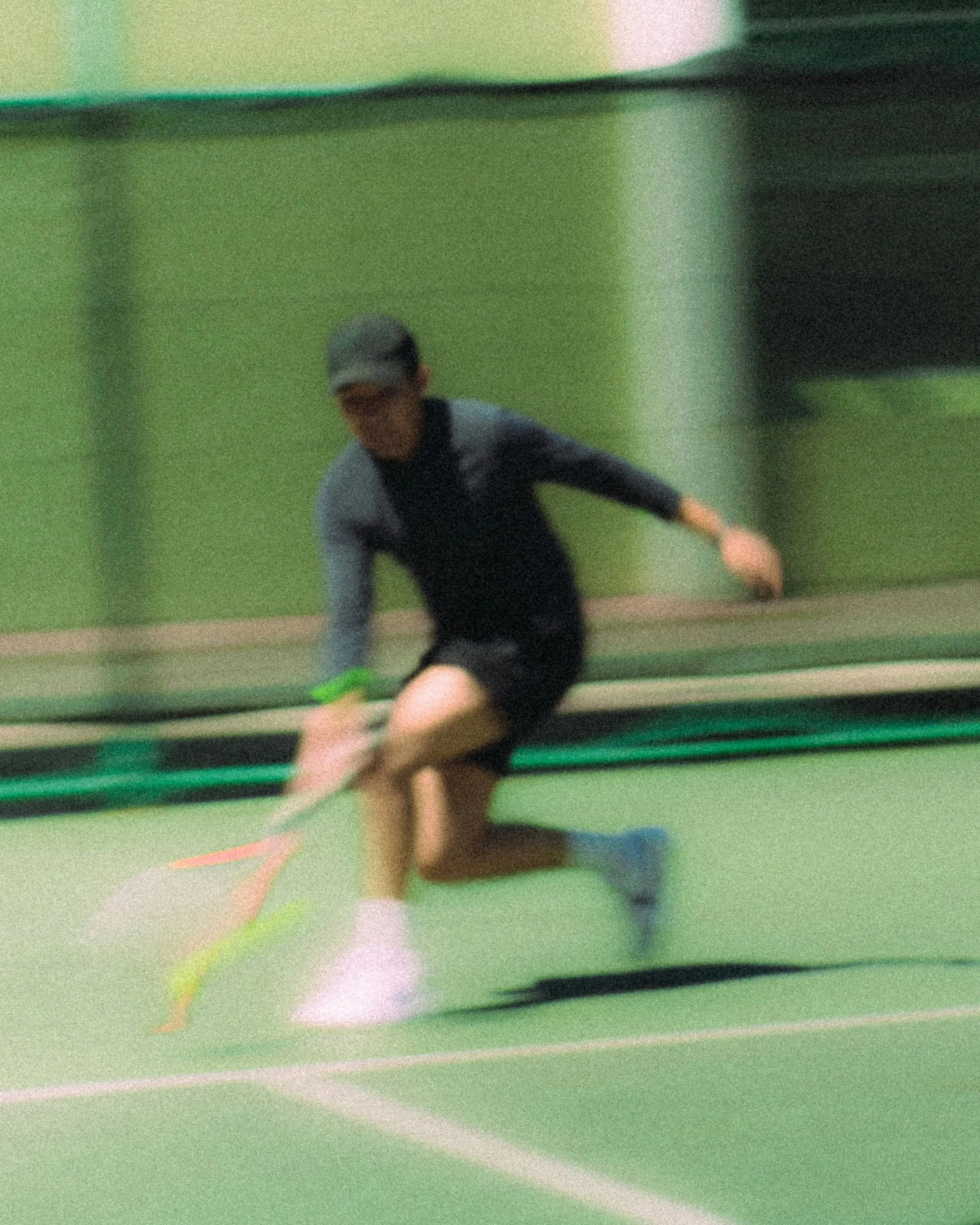 Motion-blurred photograph of a tennis player lunging for a low shot on a green indoor court.