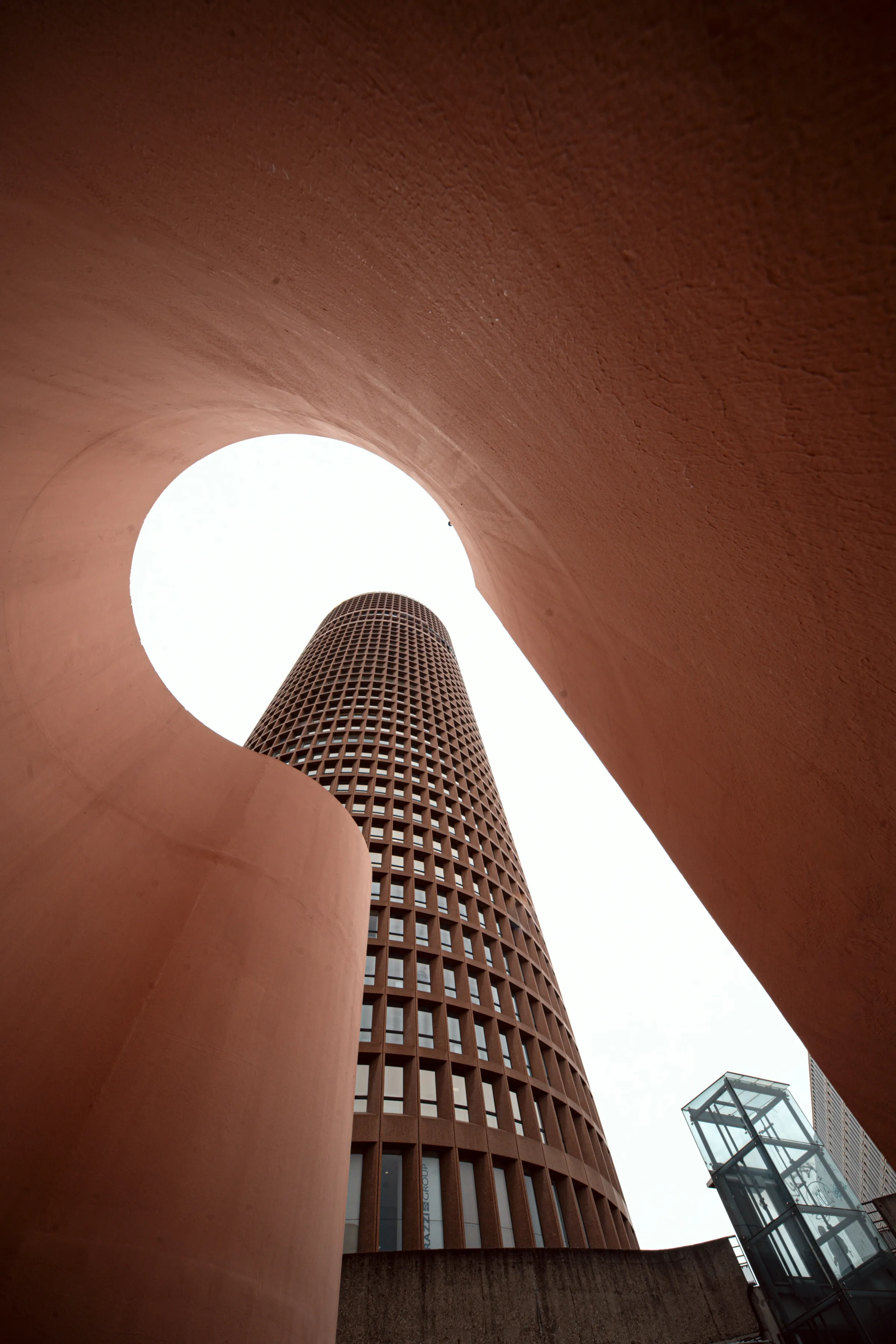 Upward view of a cylindrical terracotta-red office tower framed through a keyhole-shaped opening in a curved salmon wall.