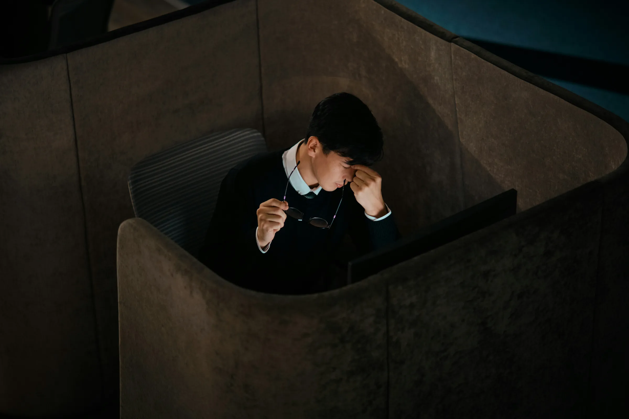 Overhead view of a man in a dark sweater rubbing the bridge of his nose while holding his glasses, seated in a felt privacy pod lit by a screen.