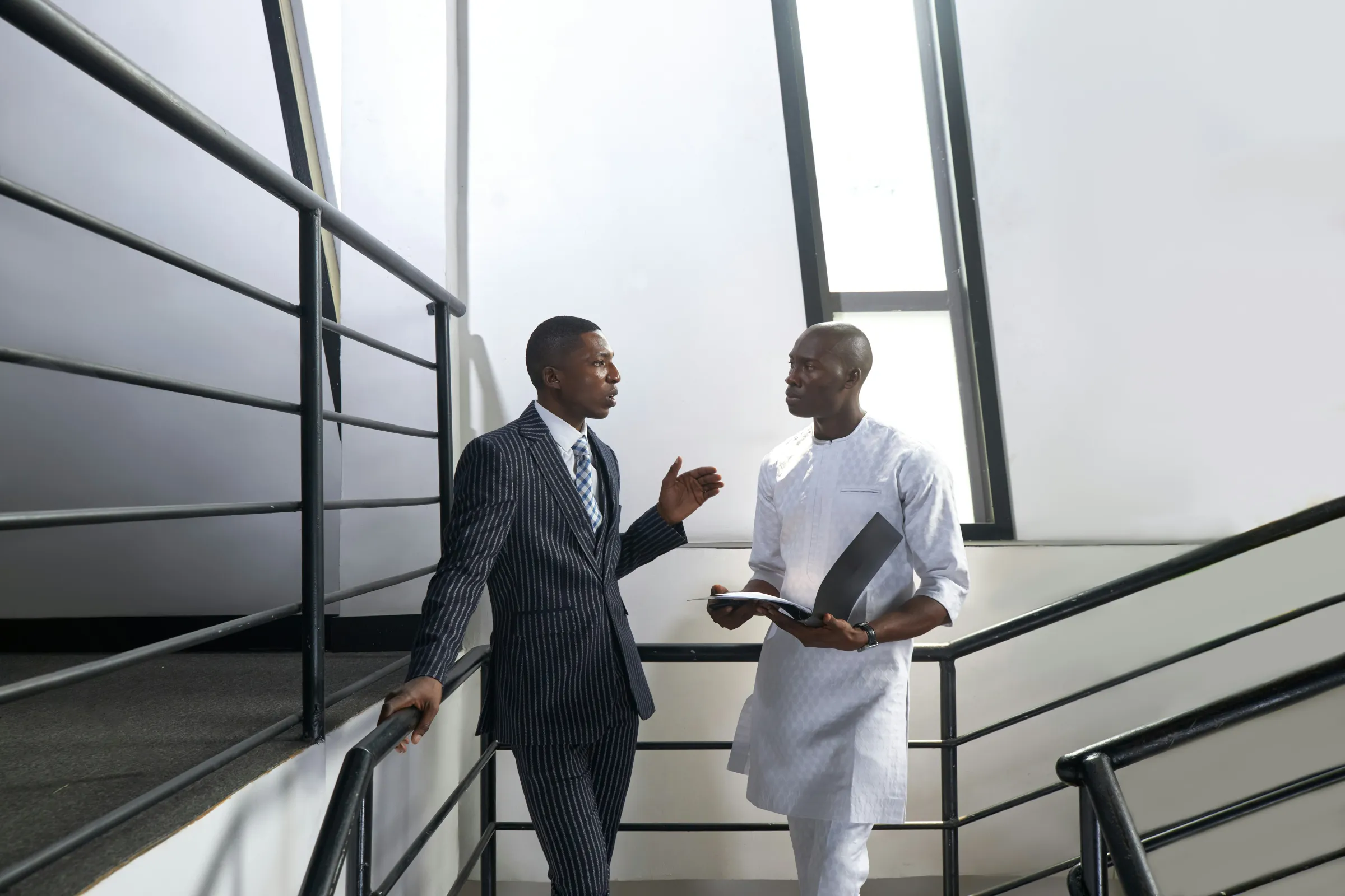 Two men in business dress, one in a pinstripe suit and one in white traditional attire, talk on a landing beside a black metal stair rail.