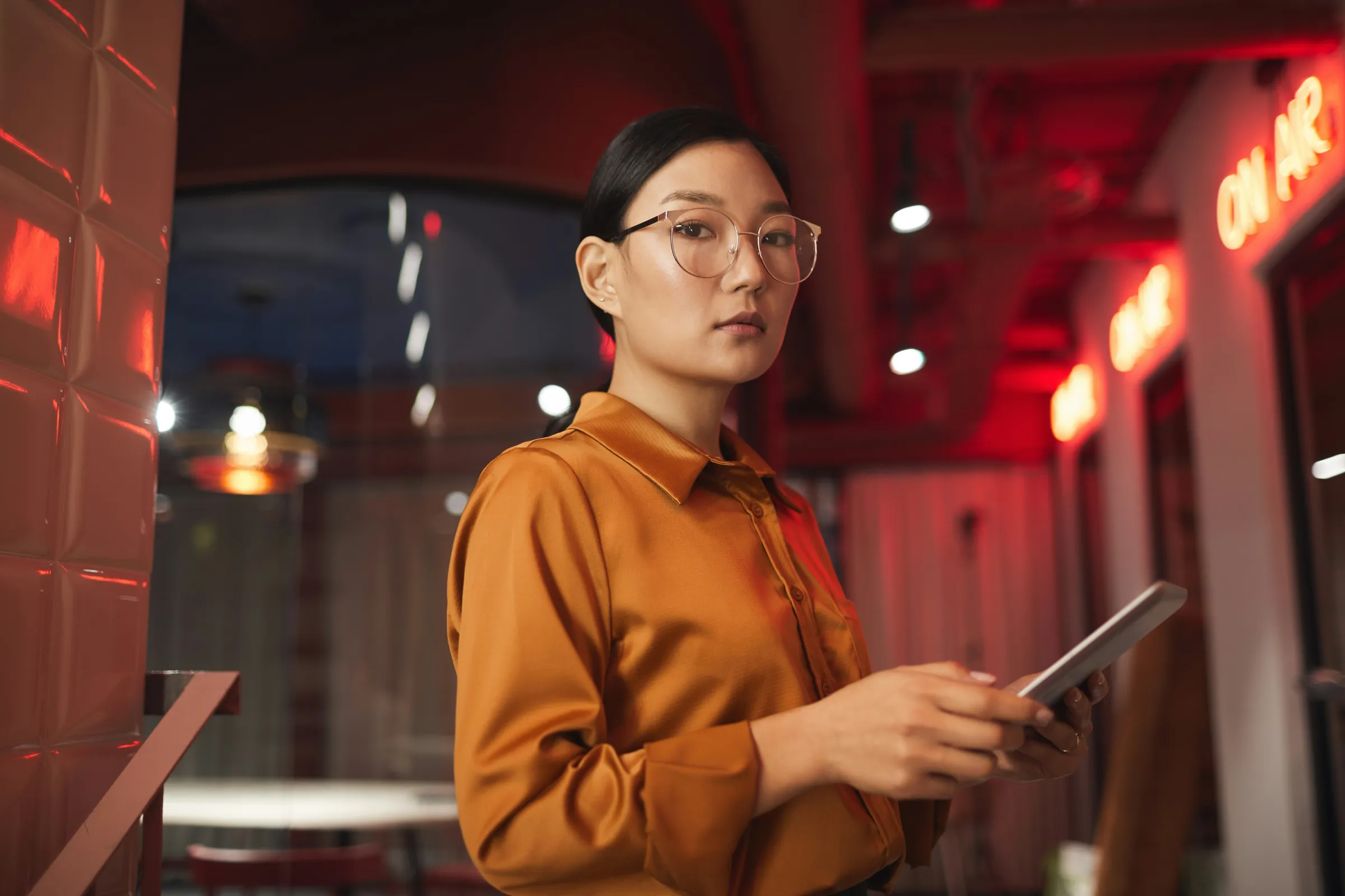 A woman in glasses and an orange satin shirt holds a tablet in a dark bar lit by red 'On Air' neon signs.